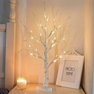 Lampe de chevet LED arbre blanc, allumée et posée sur une table de chevet à côté d'une bougie allumée et d'un cadre blanc.