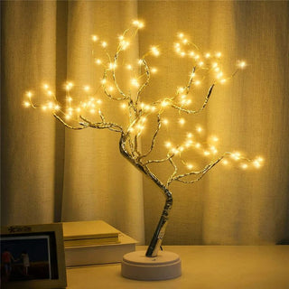 Lampe de chevet LED arbre, allumée et posée sur un meuble, derrière on voit un rideau et à côté deux livres posées l'un sur l'autre.