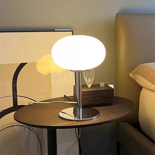 Lampe de chevet post-moderne et minimaliste en verre, allumée et posée sur une table de chevet ovale en bois foncé. Derrière, un grand cadre est posé au sol et contre le mur.