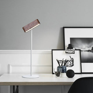 Lampe à poser bois aimantée sur pied métallique, allumée et posée sur un bureau blanc, avec des cadres photo posés à côté de la lampe et un pot à crayon devant. Un mur gris et blanc avec des moulures derrière. Tout est très moderne.