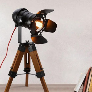 Lampe à poser bois et métal noir style projecteur vintage, allumée et posée sur un bureau en bois à côté de livres.