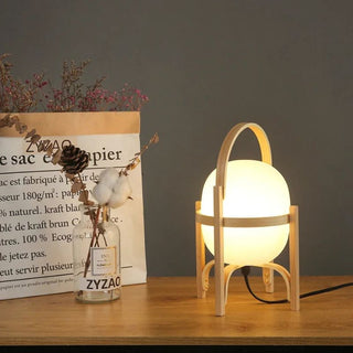 Lampe à poser bois LED en verre et bois au design moderne, allumée et posée sur un meuble en bois, à côté d'un vase en verre avec des plantes séchées, derrière un mur gris foncé.