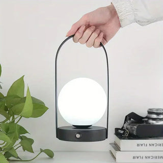 Lampe à poser sans fil LED portative à intensité variable dans la main d'une personne avec une plante verte à gauche et un appareil photo posé sur des livres à droite