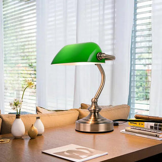 Lampe à poser vintage avec abat-jour vert installée sur un bureau avec des livres et des pots de fleur avec un canapé devant et des fenetres avec des rideaux et des stores en fond