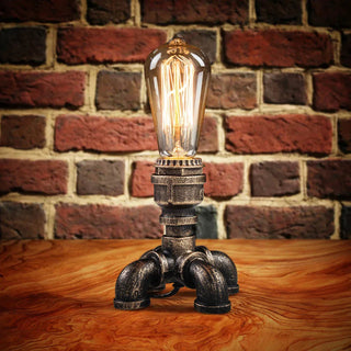 Lampe à poser vintage à LED en forme de tuyaux posée sur du bois avec un mur en brique en fond