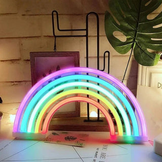 Veilleuse à poser arc-en-ciel LED style néon, allumée, posée sur un bureau, devant un objet décoration en forme de cactus en métal avec un cadre photo, et une feuille synthétique de plante verte.