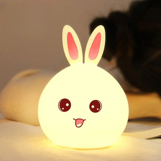 Veilleuse à poser en forme de lapin à LED allumée sur un tissu beige avec un livre à droite 