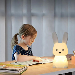 Veilleuse à poser lapin tactile 7 couleurs, posée sur un bureau en bois, une petite fille lit un livre