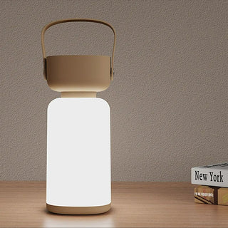 Veilleuse à poser LED portable de camping, beige et blanche, avec une anse posée sur une table en bois avec deux livres à côté d'elle.